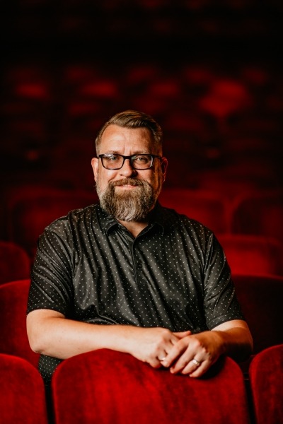 man with brown hair with beard and glasses, grey shirt sitting in red theater seats