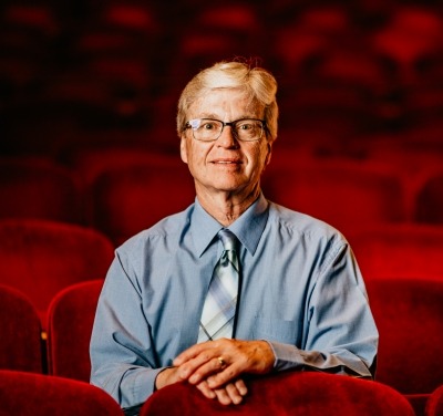 Man with white hair, black glasses, blue button up and tie sitting in red theater seats