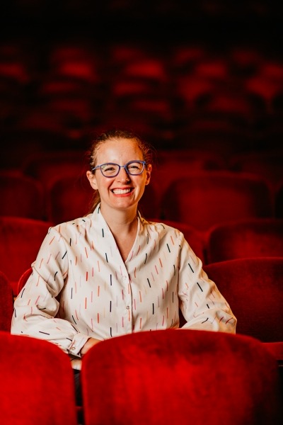 woman with brown hair, blue glasses, multi colored blouse sitting in red theater seats