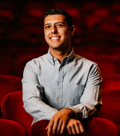 Man in blue button up with black hair sitting in red theater seats