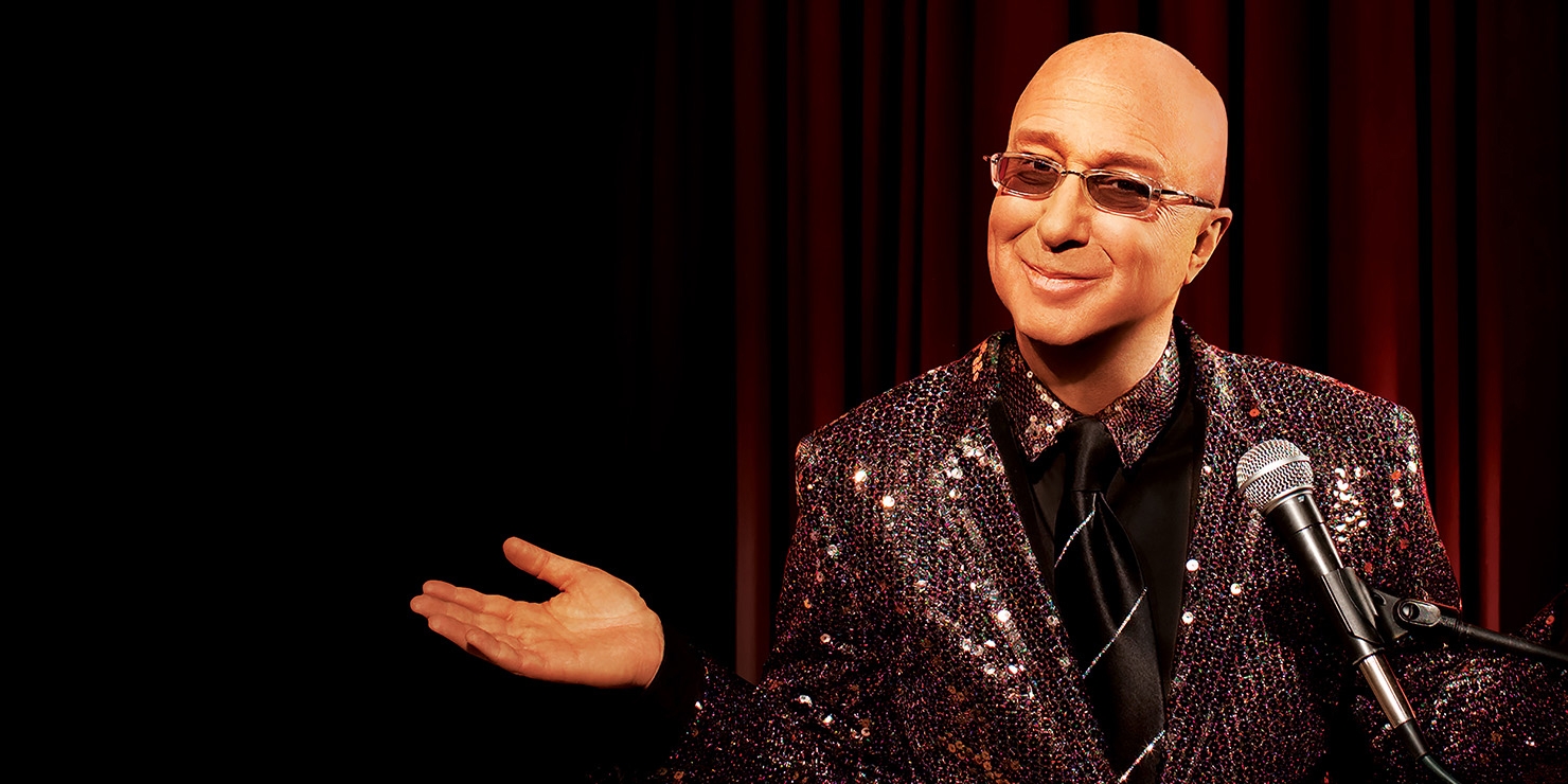 Paul Shaffer standing at a microphone wearing his signature glasses in front of a stage curtain