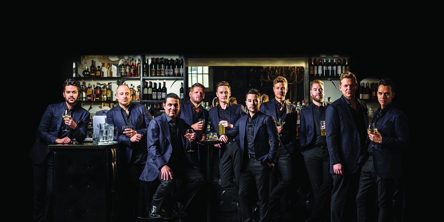 The TEN Tenors standing in line in front of a bar dressed all in black, several are holding glasses of beer or wine.