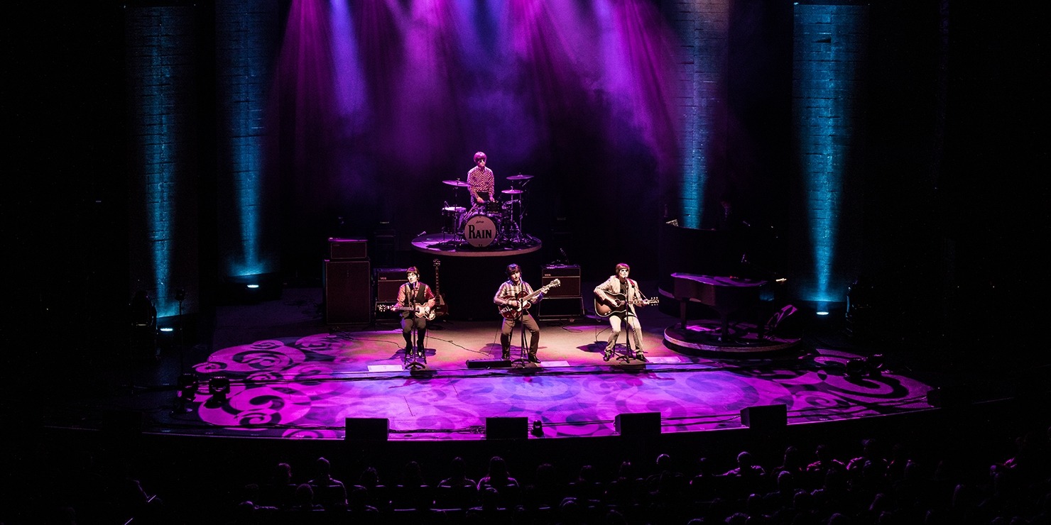 Image of the four performers in RAIN playing music in front of a live audience on a stage decorated with purple lights.