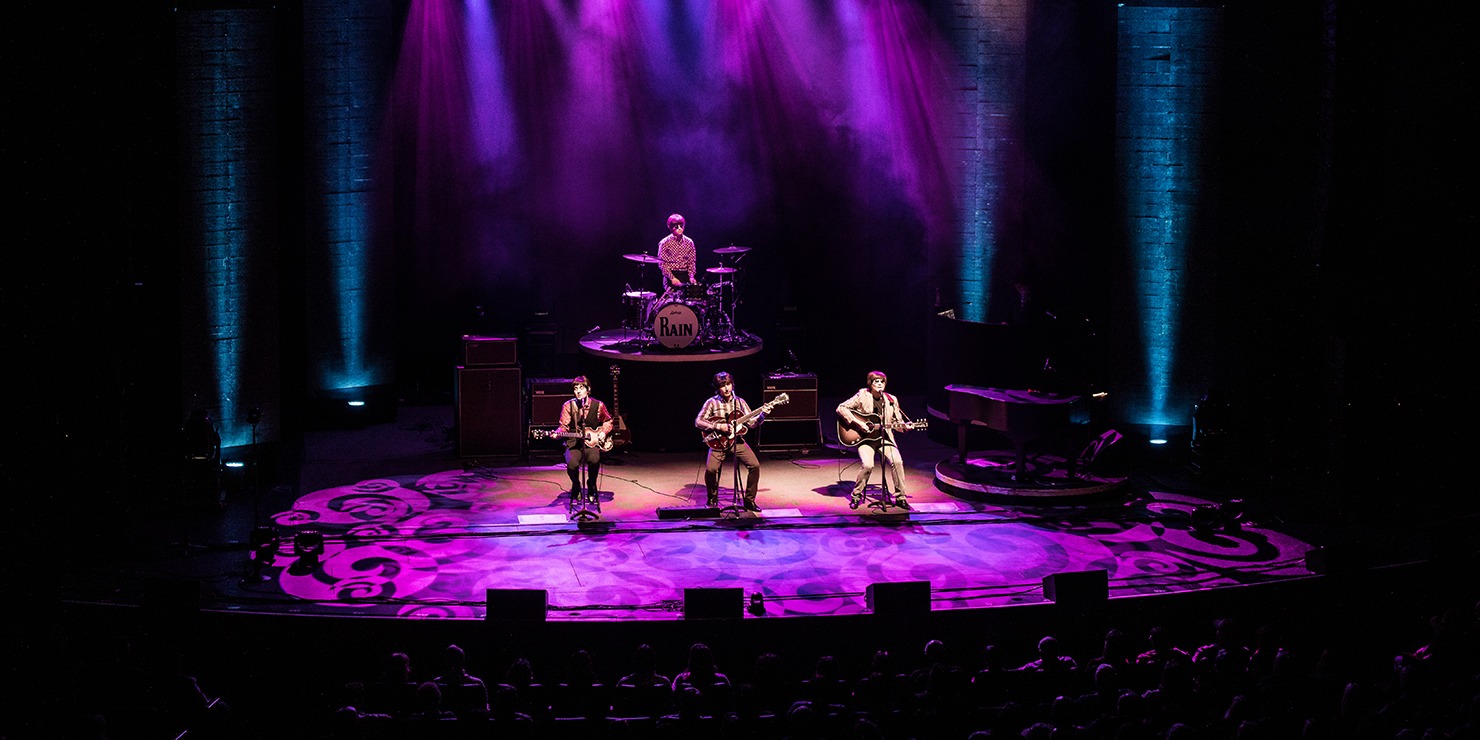 Image of the four performers in RAIN playing music in front of a live audience on a stage decorated with purple lights.