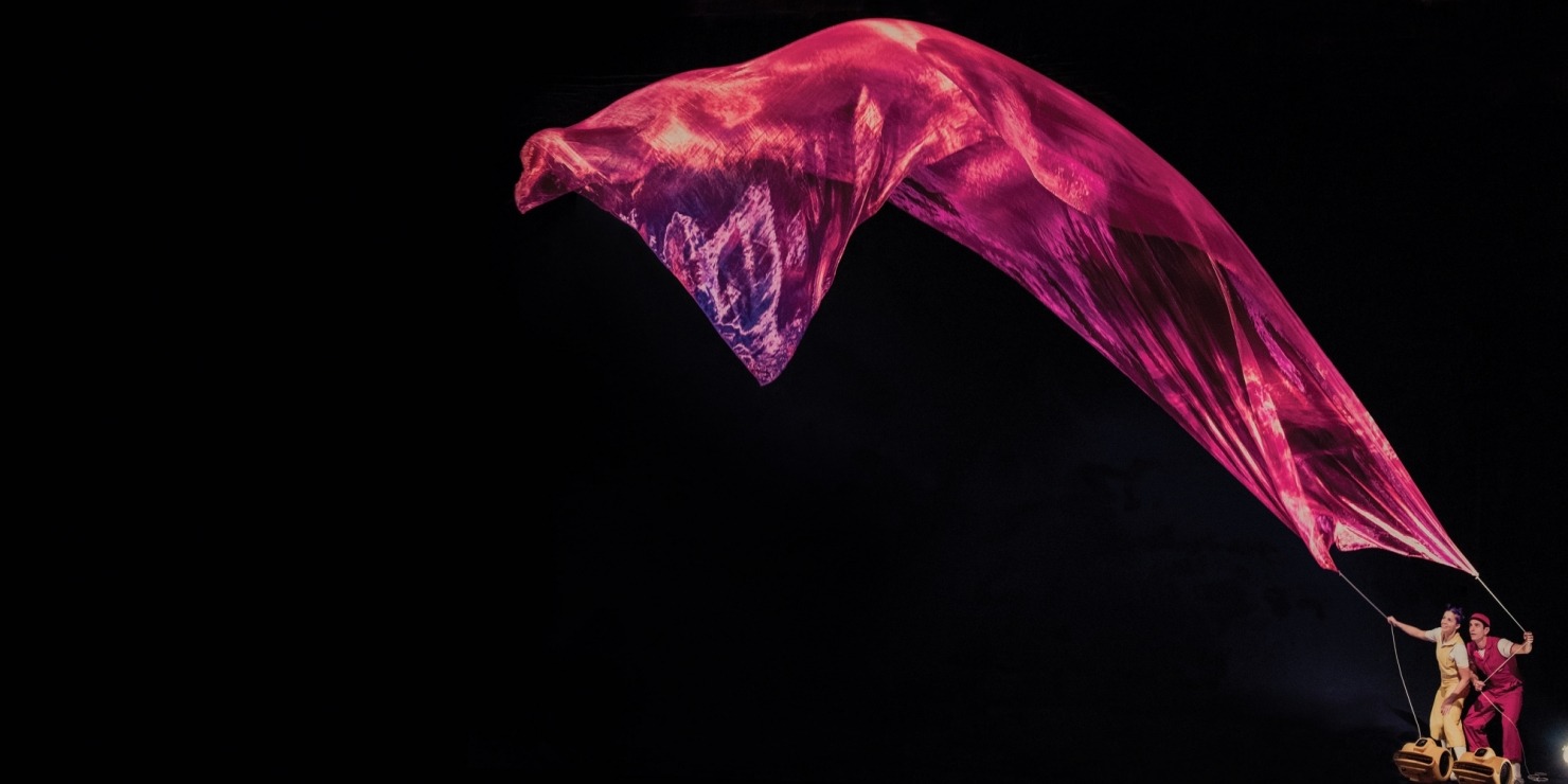 A woman in yellow and a man in red holding a string connected to a red piece of sheer fabric that's floating in front of a black background.