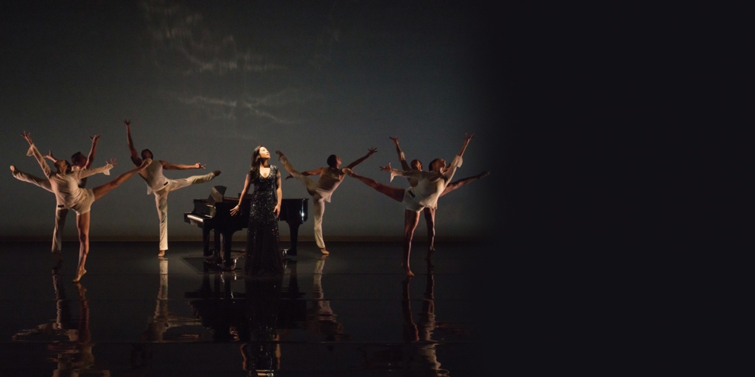 Woman in black, sparkly dress looking up standing in front of a piano. She is surrounded by six dancers dressed in white, all in front of a black background.