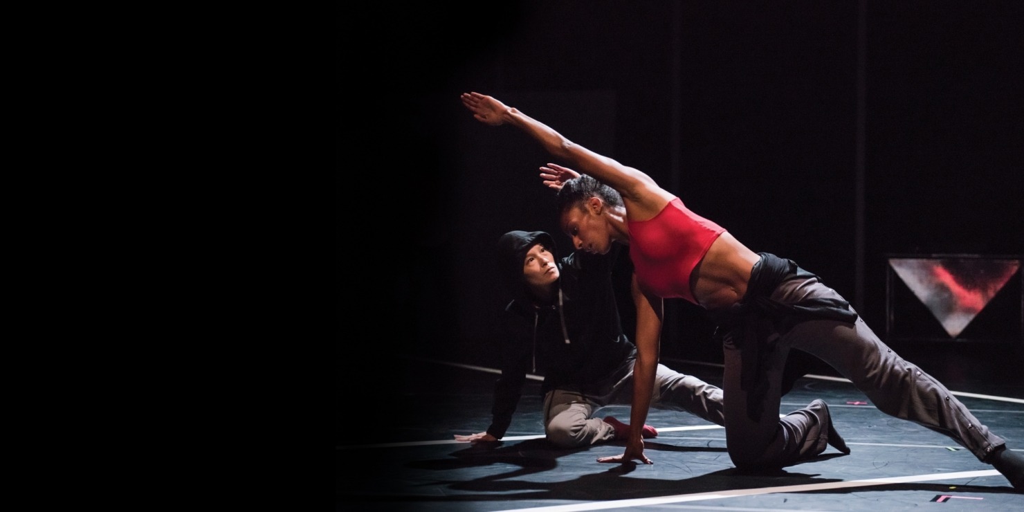 Woman in red top and grey pants on one hand and one knee reaching over her head with a person in black behind her mimicking her all in front of a black background.