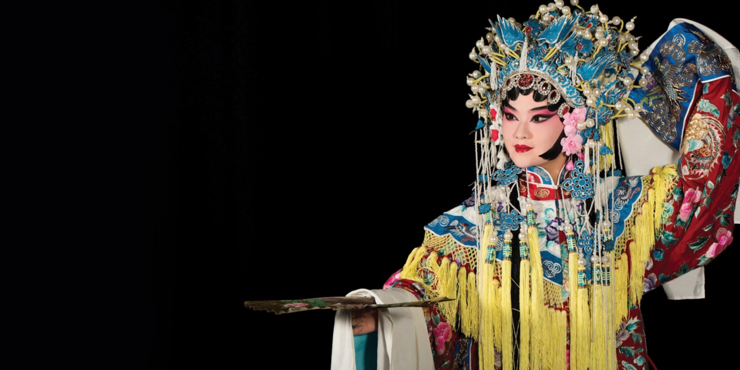 Woman in blue, yellow, and red beaded Chinese attire holding a fan in front of a black background.