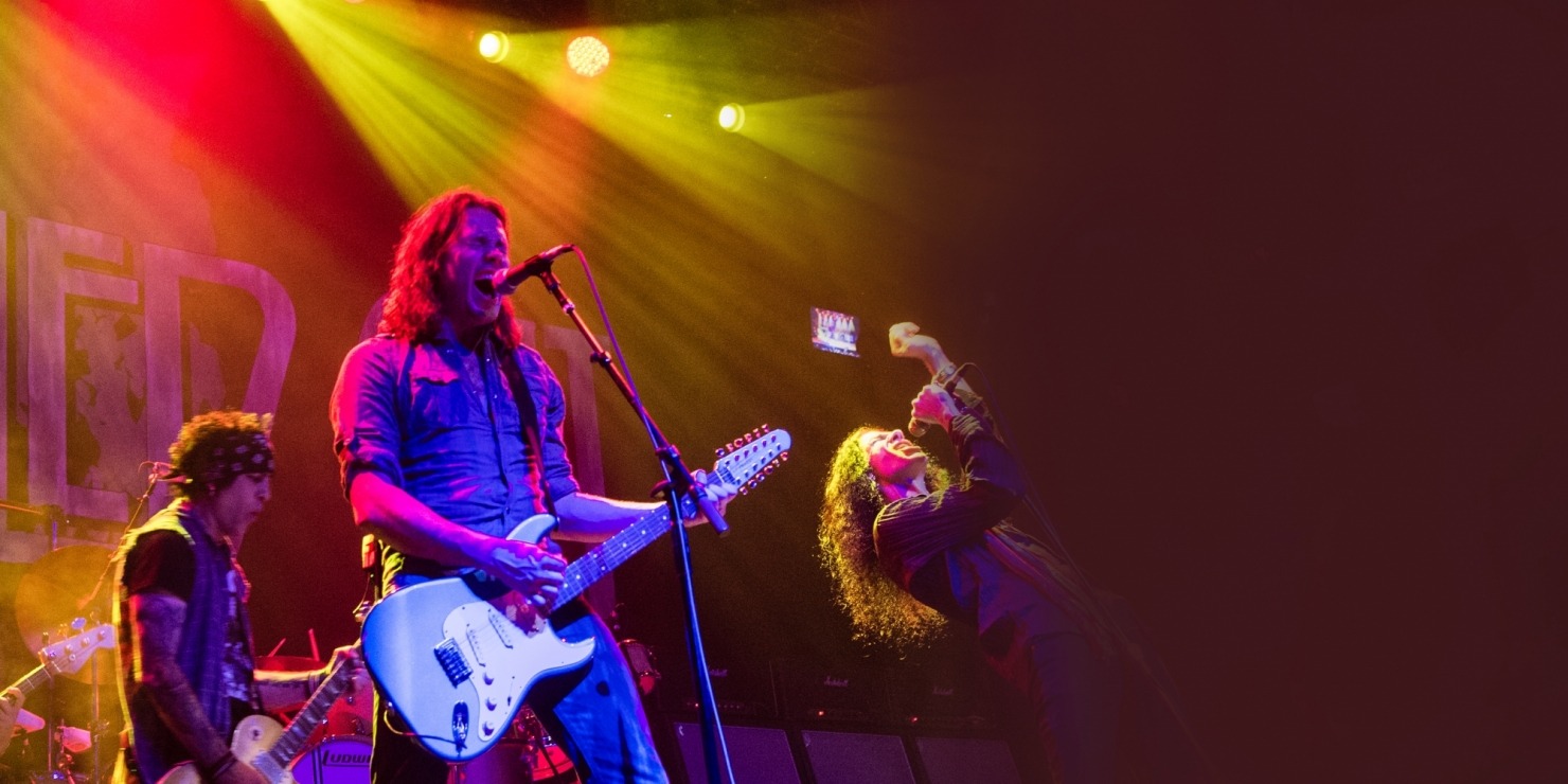 Two men playing guitar and one man singing dressed as Led Zeppelin band members in front of a pink, red, orange, and yellow background.
