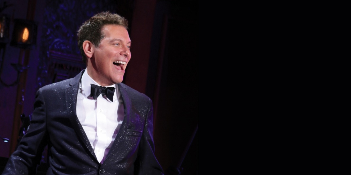 Man in a sparkling black tuxedo, white button down shirt and black bow tie smiling in front of a dark blue, purple and red background.