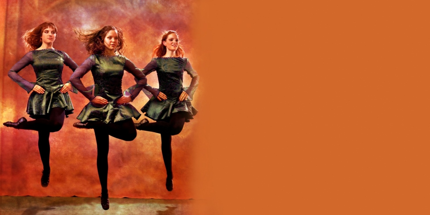 Three women Irish dancing in green dresses and black tights in front of an orange background.