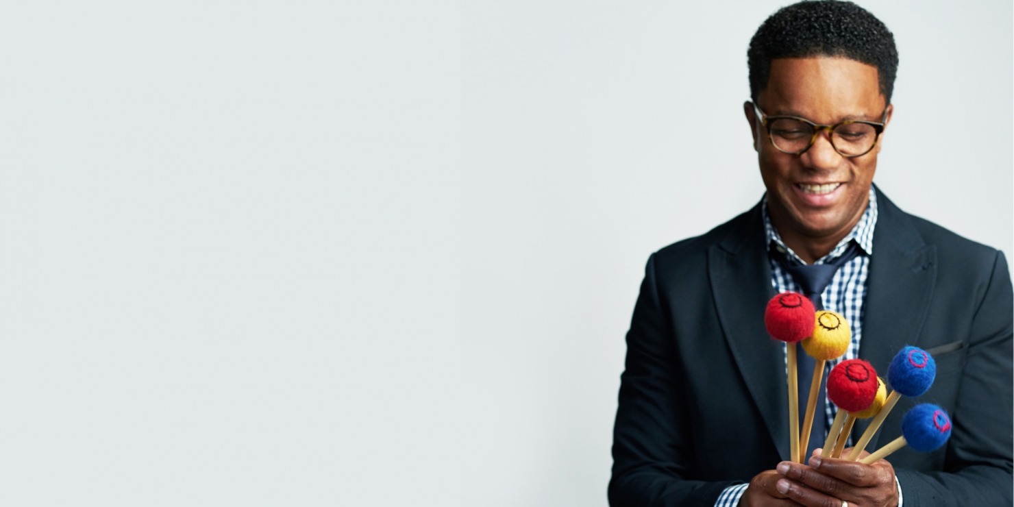 Man with glasses in a black tux, gingham button down and black tie holding red, blue and yellow vibraphone mallets in front of a light grey background.