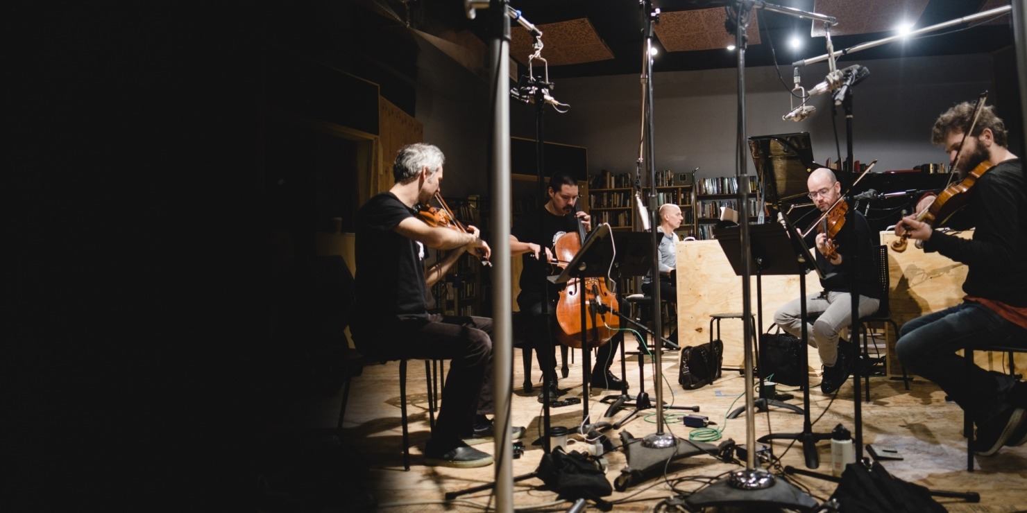 Five men sitting in a recording studio playing stringed instruments in front of a grey wall
