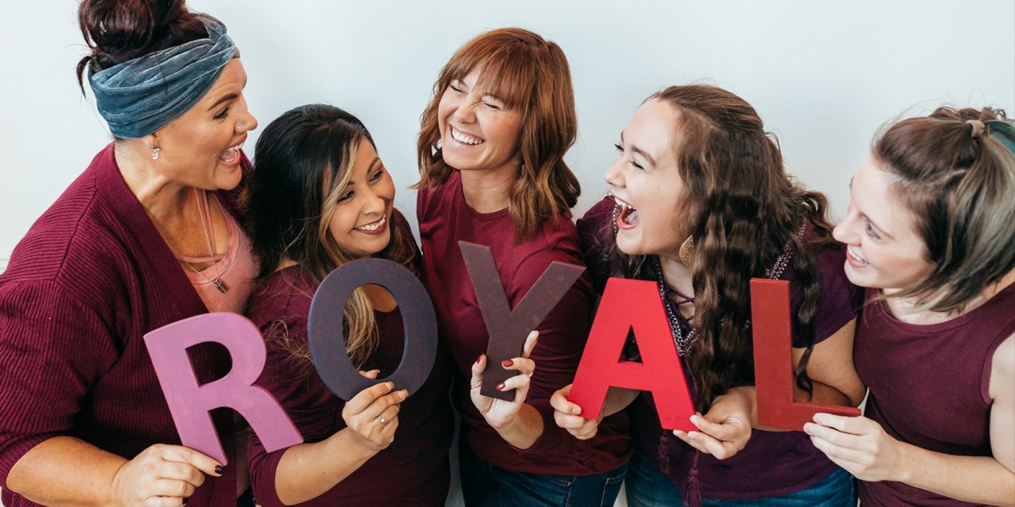 5 Women wearing purple laughing holding letters that spell out "ROYAL" in front of a light grey background.