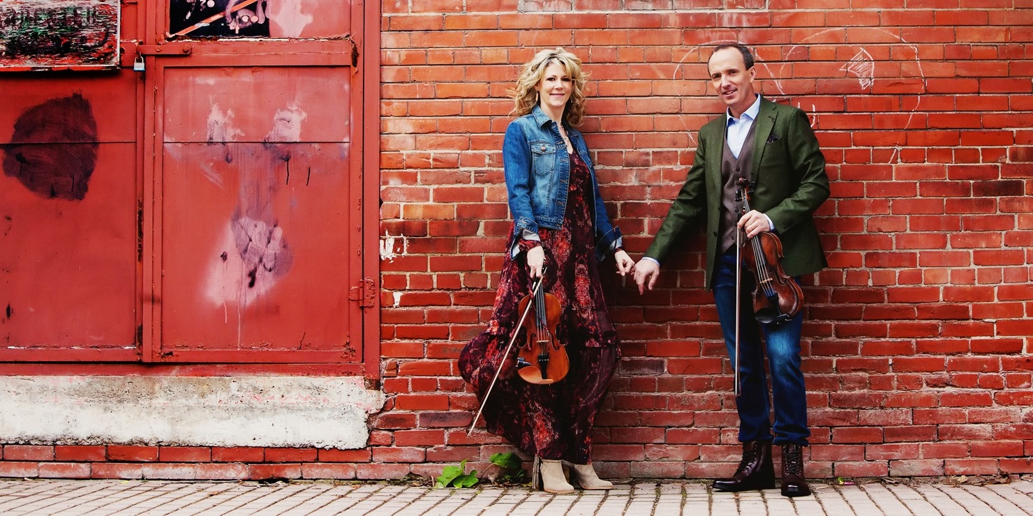 Image of the two performers from Celtic Celebration holding hands and their fiddles while standing in front of a brick wall.