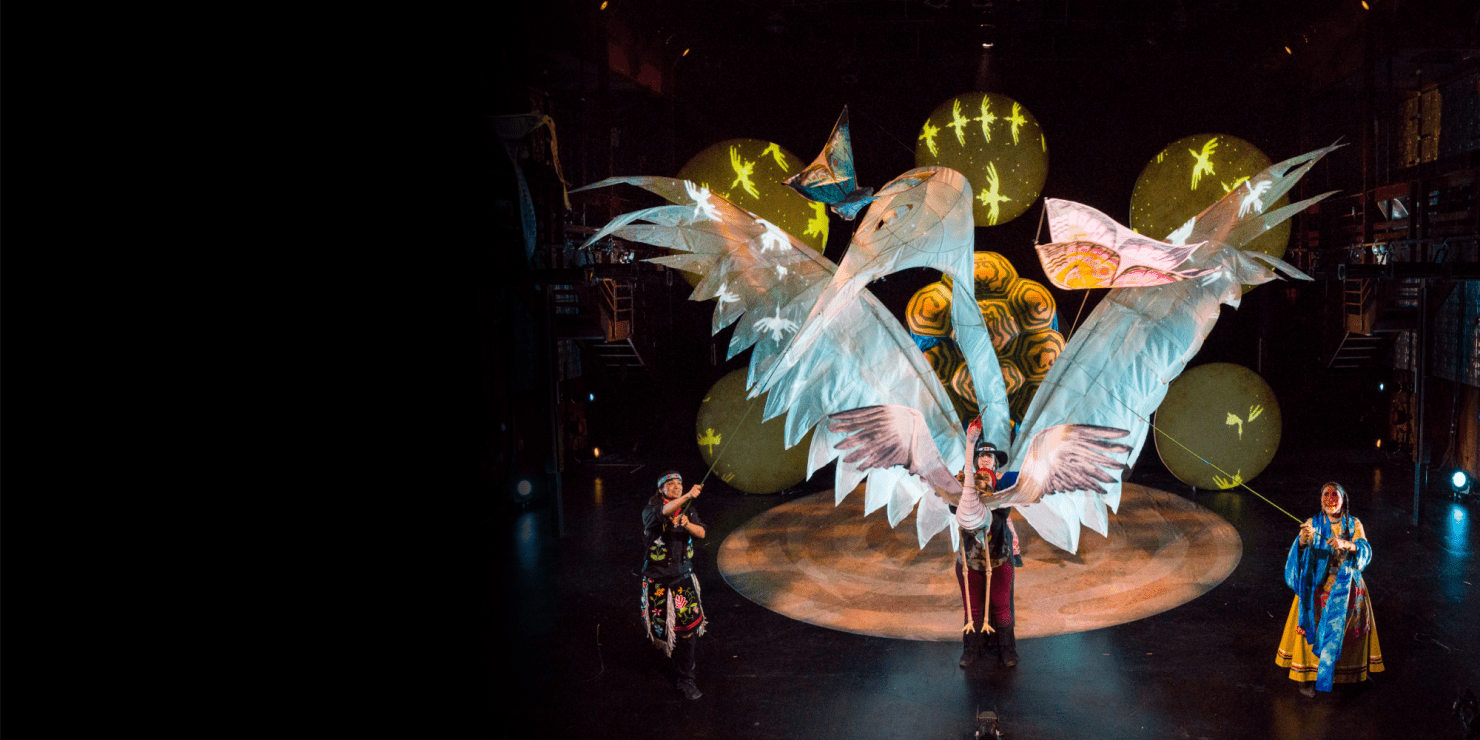 Large white crane puppet on stage. Two people holding the puppet up with strings. One person holding a smaller crane puppet at the front of the stage. Other puppets are showcased on the stage and lights are on the back wall of stage.