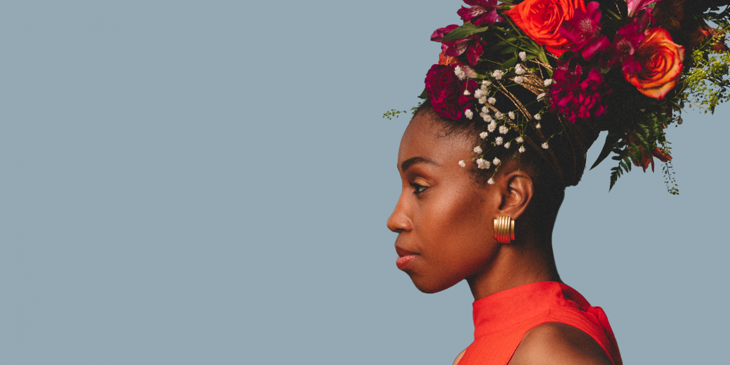 Picture of women with red dress on looking away from the photo. She is wearing a tall hat of multiple different flowers.