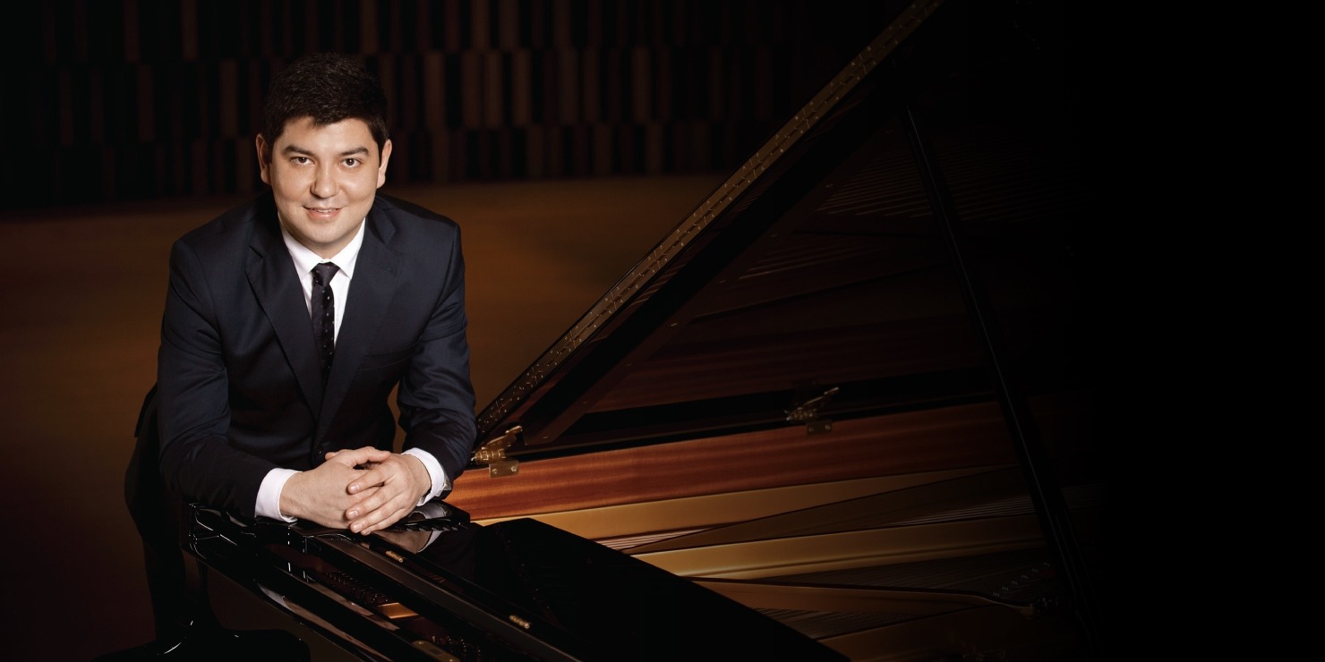 Man with dark hair in a suit and tie leaning forward onto a piano with his hands together.