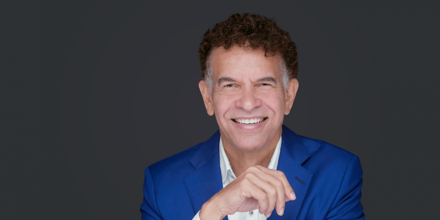 Man in blue suit jacket with white undershirt is smiling facing toward the camera. The background is grey.