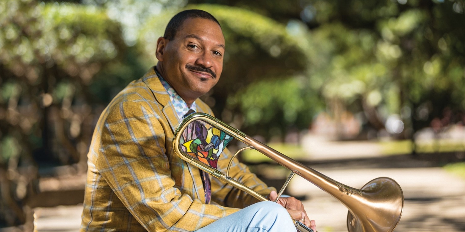 Delfeayo Marsalis in a gold jacket sitting in front of some green trees with his trombone.
