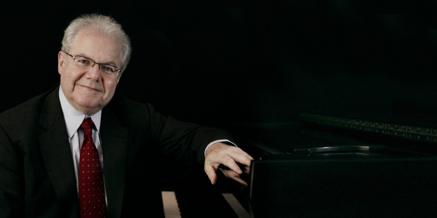 Male with white hair in suit with a red tie leaning on a piano with a black background.