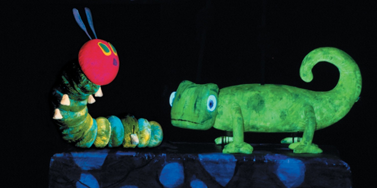Green caterpillar puppet with a green chameleon puppet in front of a black background with a blue cloud in the sky
