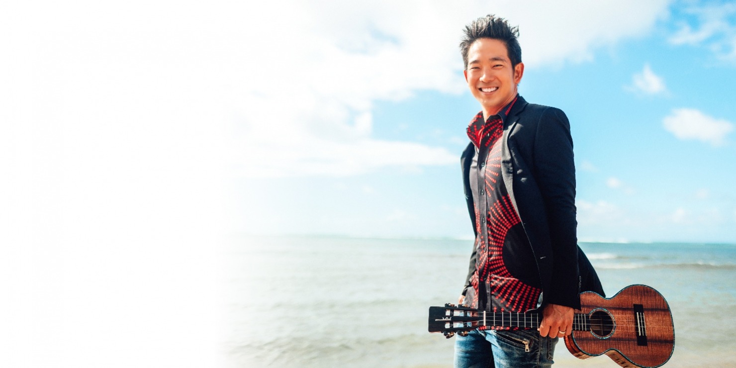 Man with dark short hair standing on a beach in a suit jacket and red and black shirt, holding a ukulele. The background shows the sky with clouds and the ocean.