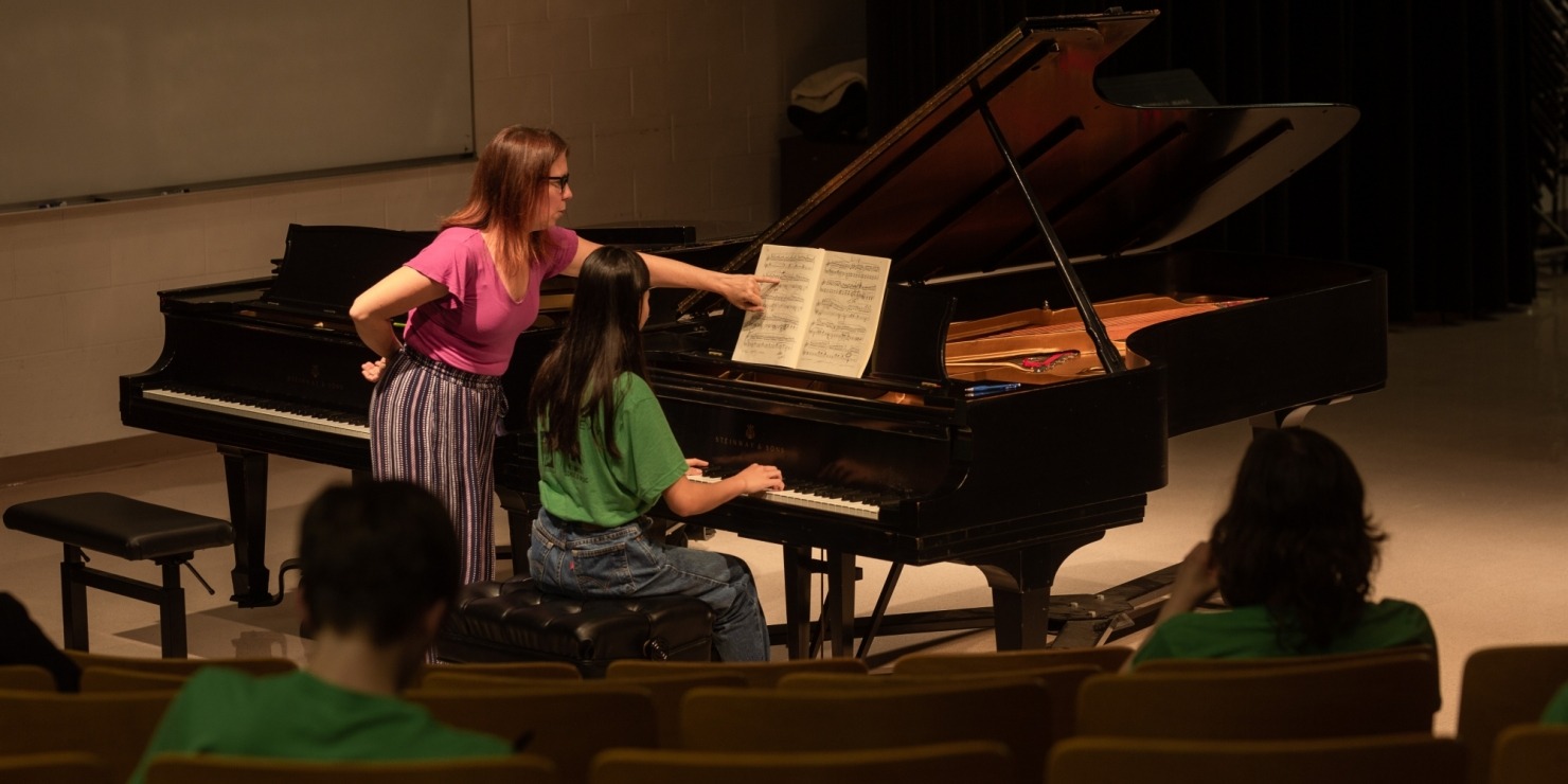 Young musician sitting and playing the piano, while a teacher is a pink shirt is helping teach.