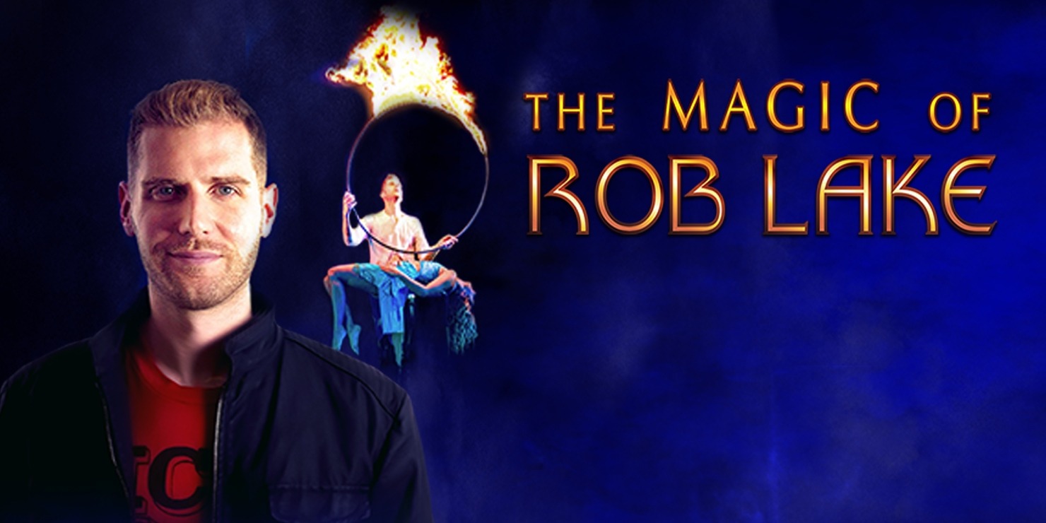 Man with a red shirt and black jacket looking at the camera. The man has short hair. Behind the man is a man holding a ring that is on fire with a lady on her back facing the sky in a blue outfit underneath the ring of fire.