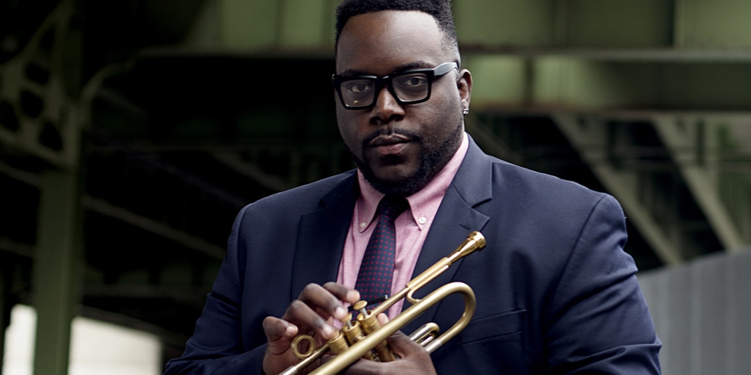 Man in a blue suit and a pink/mauve shirt holding a trumpet in front of a green and grey background.