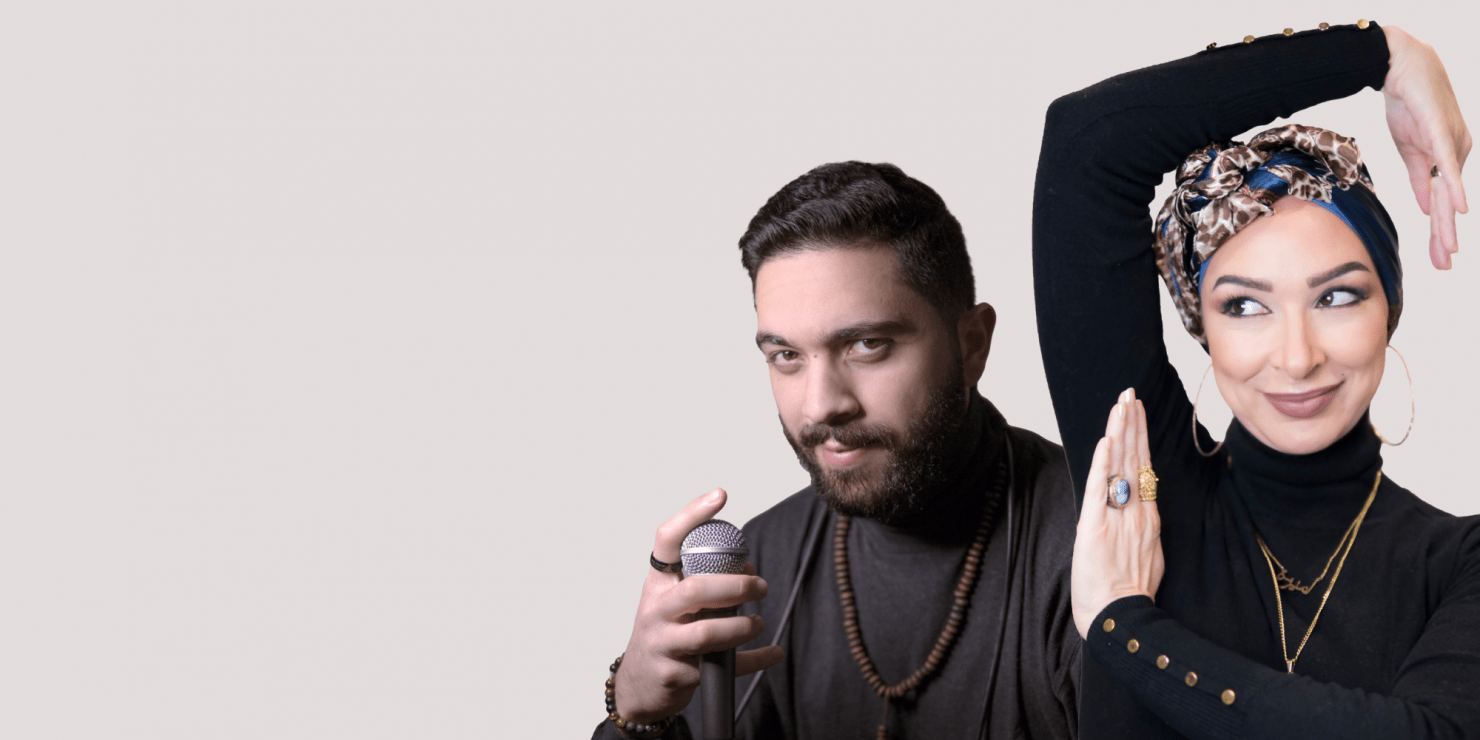 A man looks at the camera with a smirk and one eyebrow raised. He holds a mirophone in his right hand. A woman is next to him and faces the camera smiling. She has her arms raised and posed around her head. She wears gold jewelry and a black turtleneck sweater.