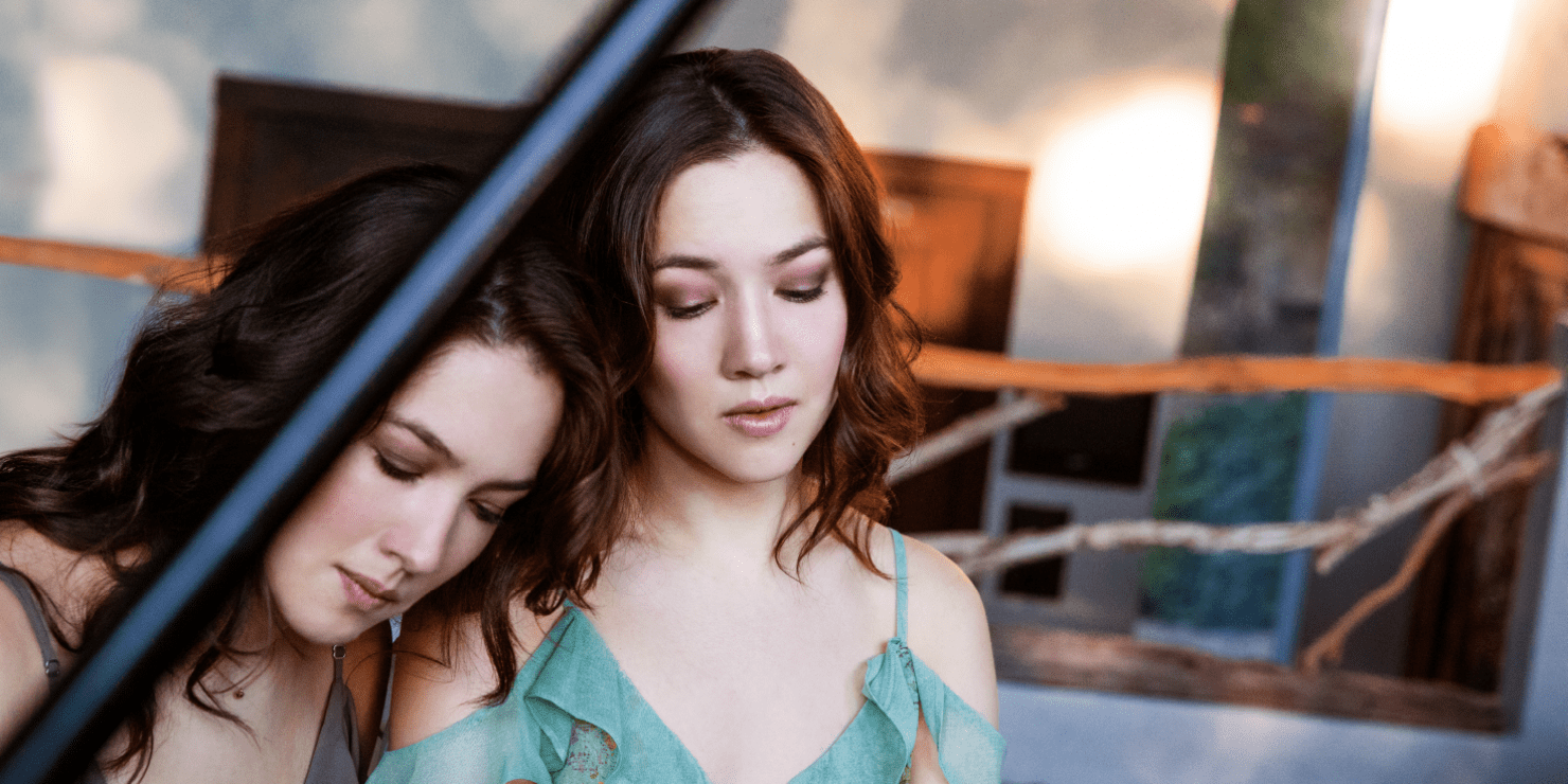 Two women with brown hair and dresses sitting next to each other. One with their head set on the other's shoulder. Both women are looking down.