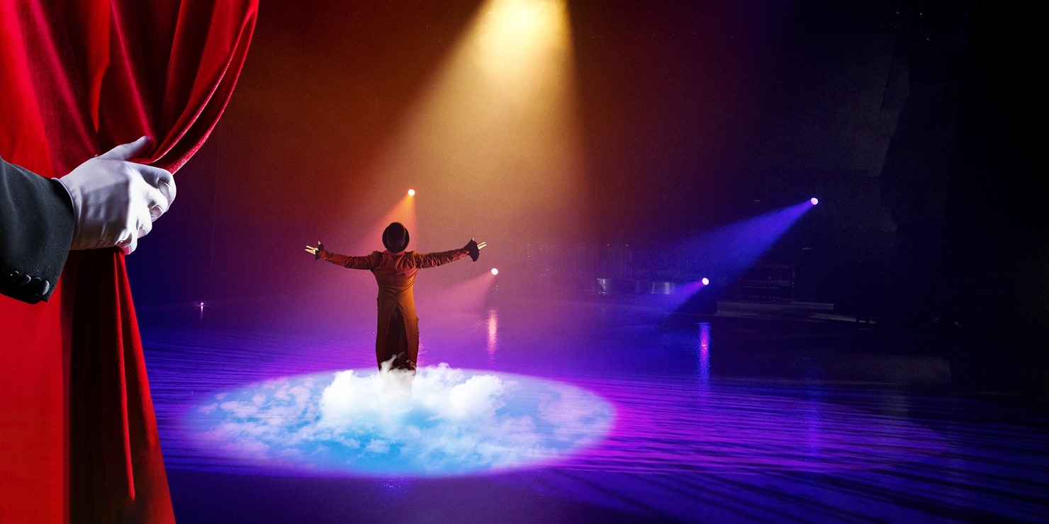 A man in a red coat in a spotlight in front of a blue background with a gloved hand pulling back a red curtain showing the man.