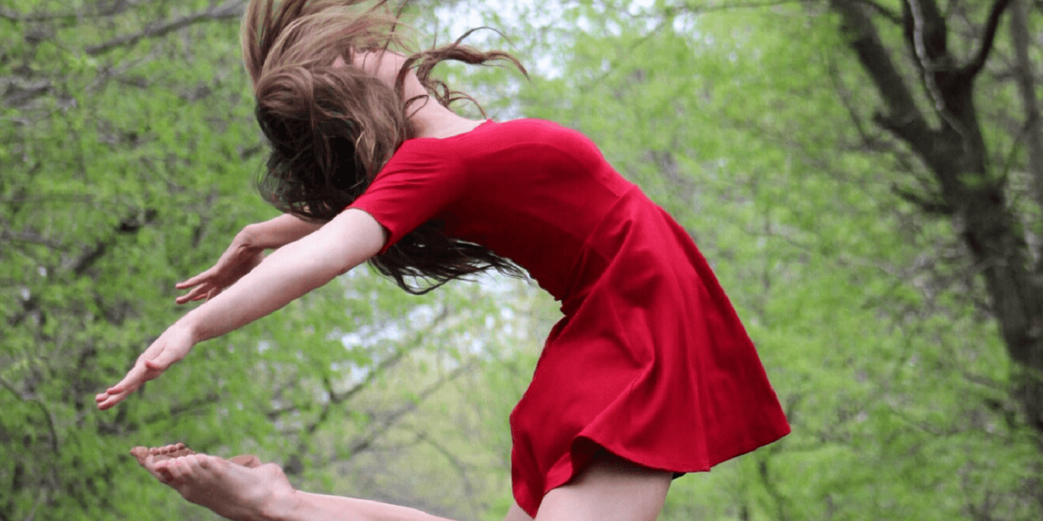 Image of female dancer in red dress. She is jumping in the air bending her arms and legs together behind her back. There is a backgound of forest trees behind her.