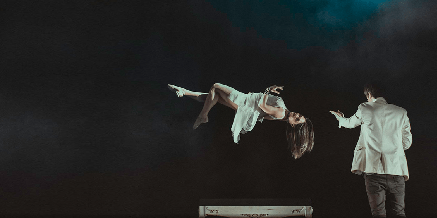 Image of women in white dress floating above a table. Man in white suit jacket is pointing arm towards floating woman, suggesting that he is directing her movement.