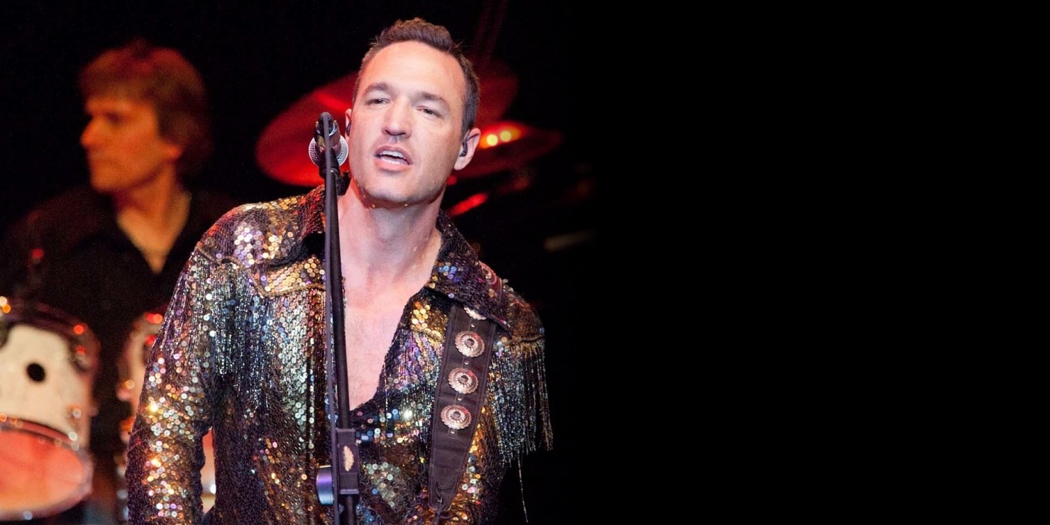 Man with dark short hair in a sparkly outfit with a guitar and a microphone singing. Member of the band is playing the guitar behind him.