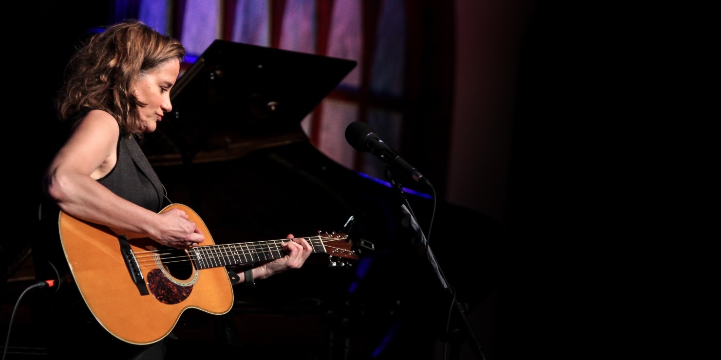 Woman with brown hair facing the right with a black sleeveless top on strumming a guitar.