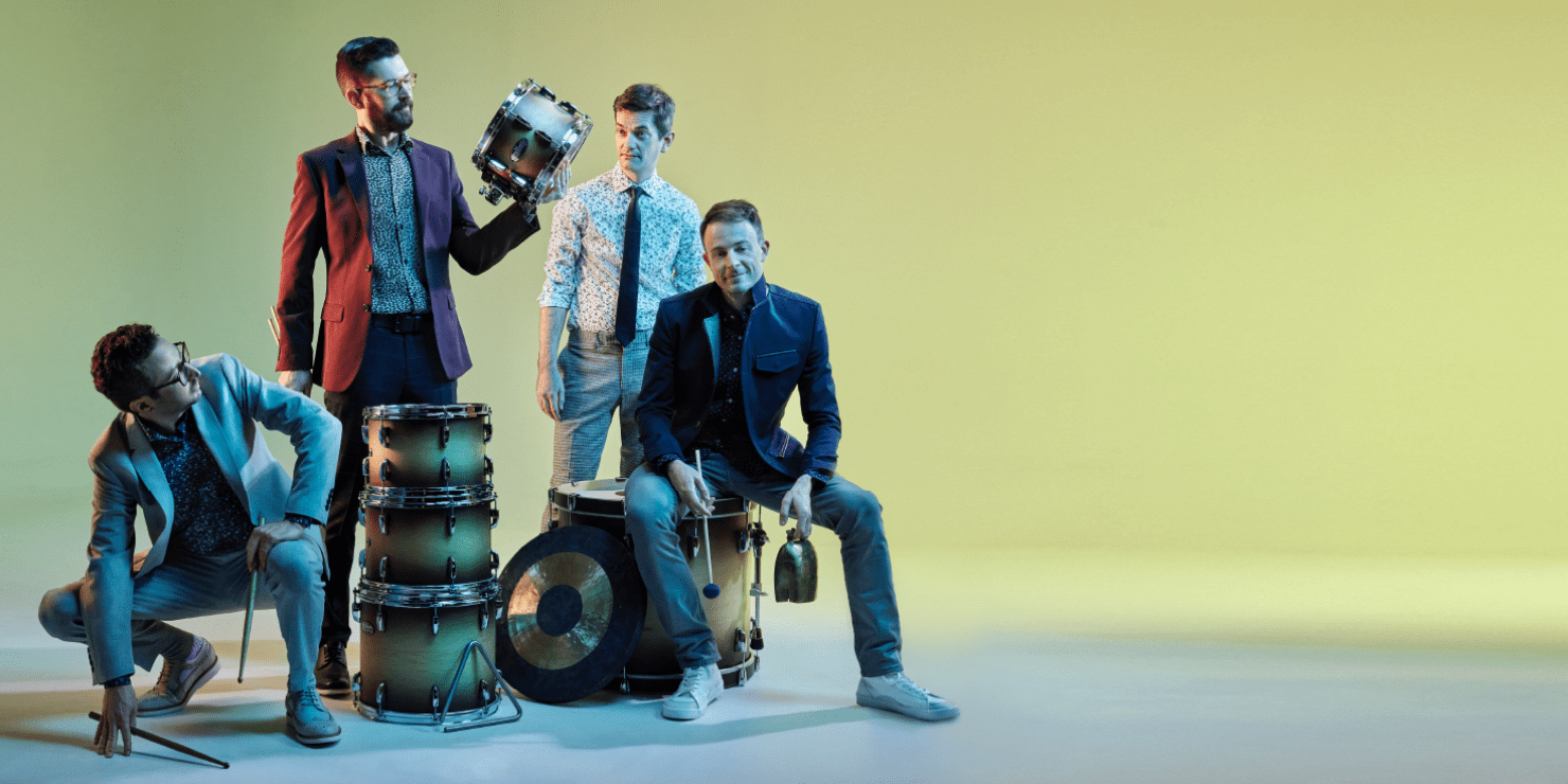 Four men in suits standing around a drum set that is not ensembled. One man is holding a cow bell. One man is holding drum sticks. One man is holding a piece of the drum set.