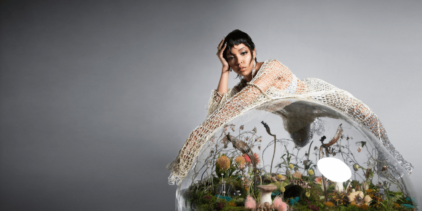 Image of a girl with short, dark hair laying across a glass ball terrarium. She is wearing a cream cover-up with a white top under it. She is looking towards he camera.