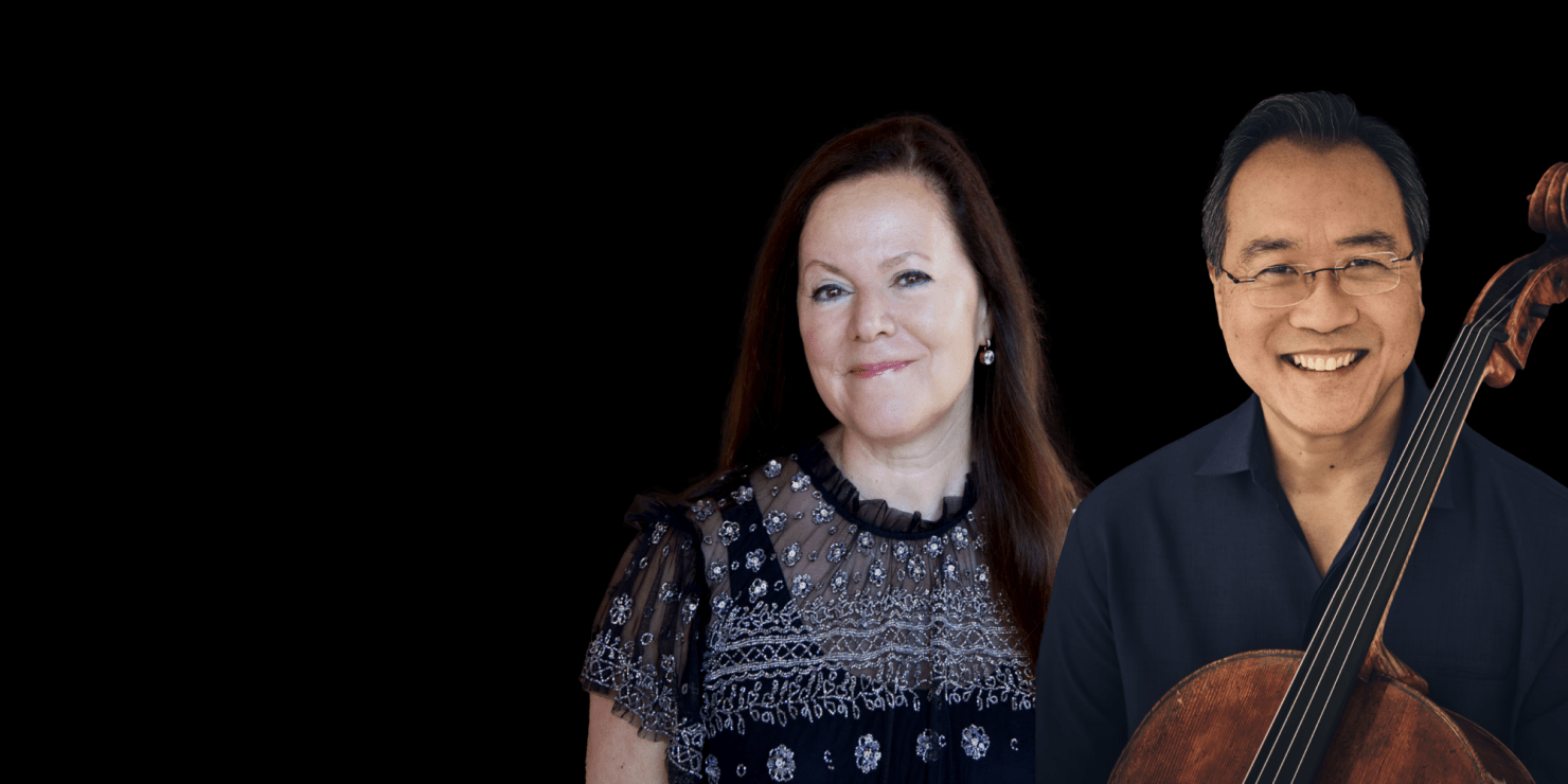A woman with brown, long hair and a man wearing eyeglasses smiles at the camera, holding his chello tightly to his chest all in front of a black background