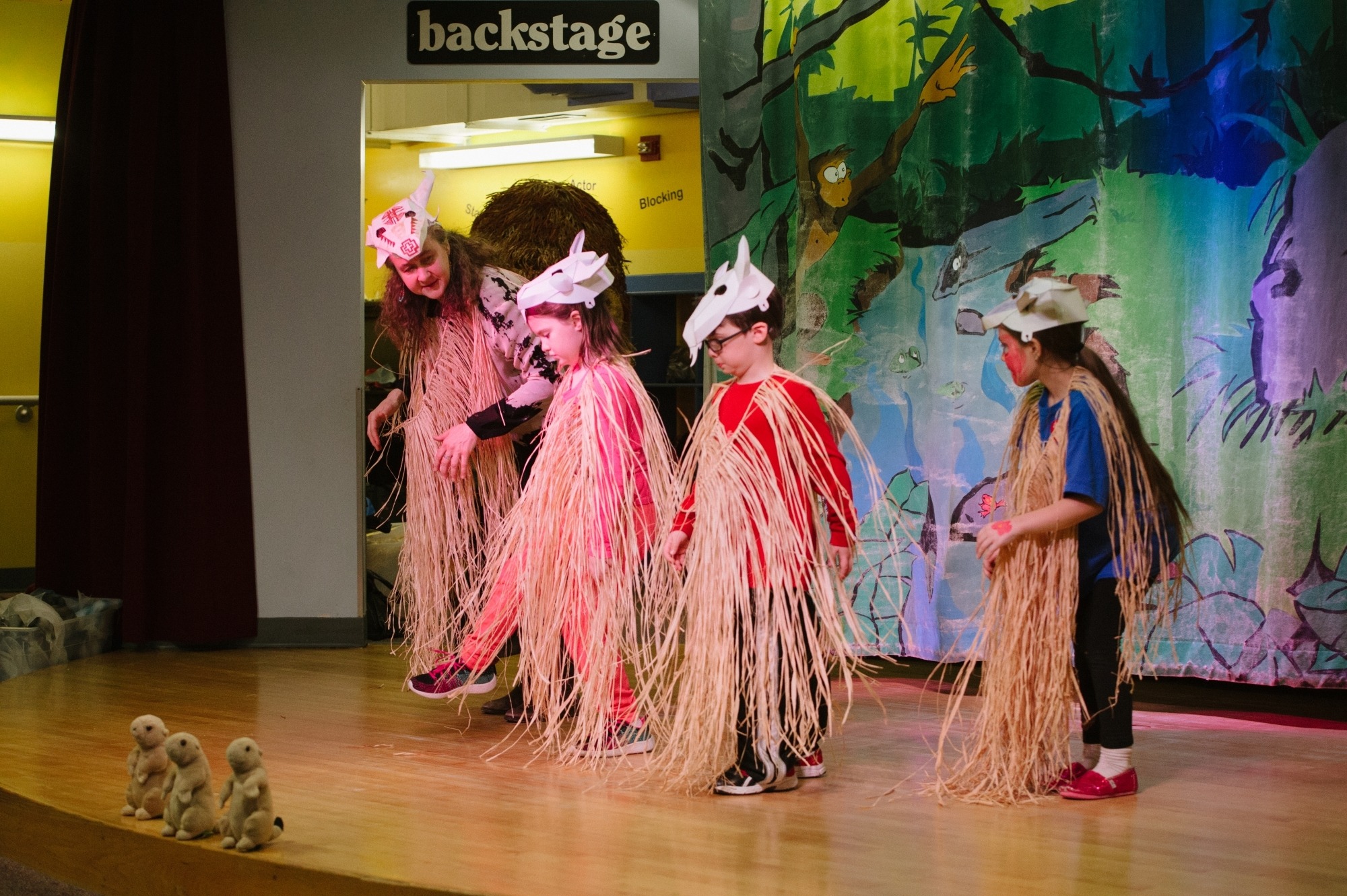 Image of three children on stage with Heather Henson wearing grass outfits and paper hats shaped like animal skulls