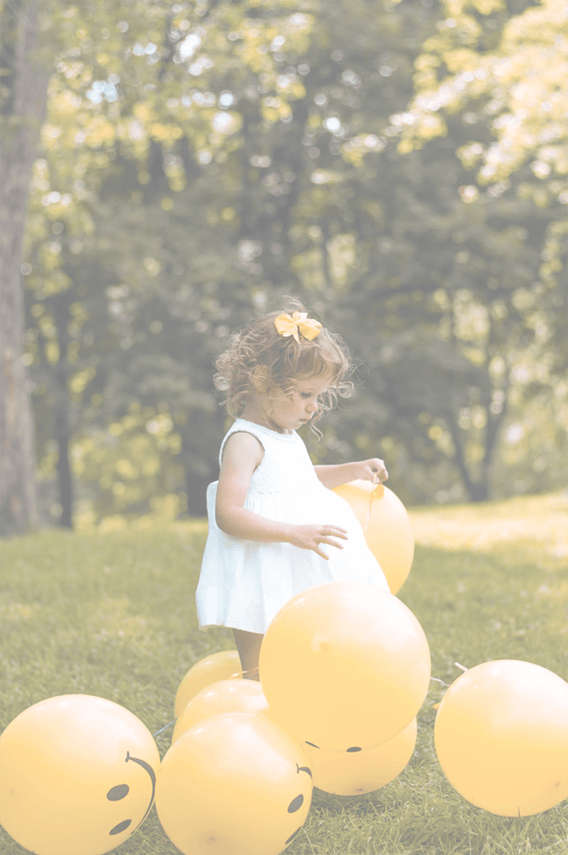 Image of a young girl in a white dress playing with yellow smiley-face balloons in the green grass