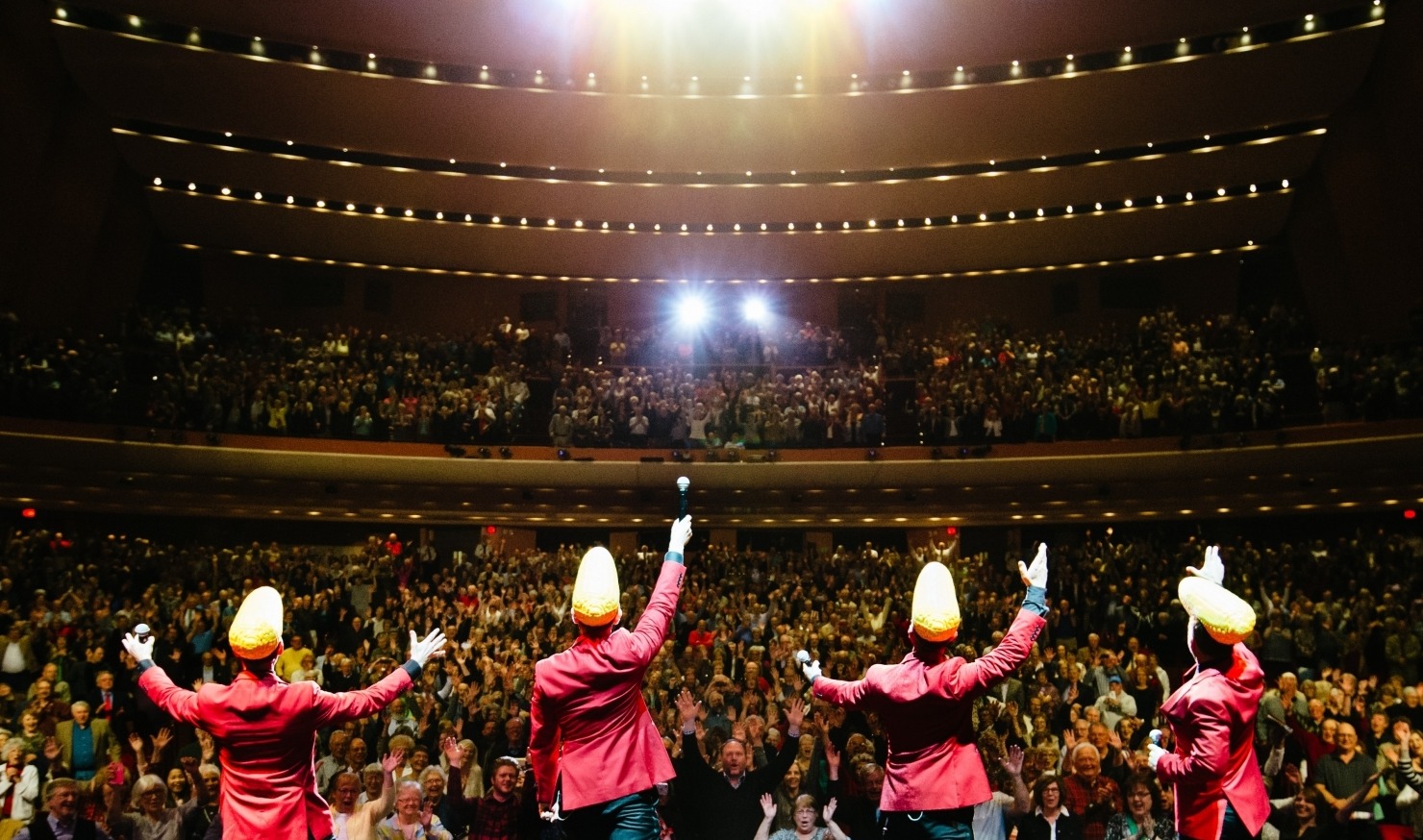 Midtown Men performing in red suit jackets with cornhead hats at the Lied Center for a packed crowd.