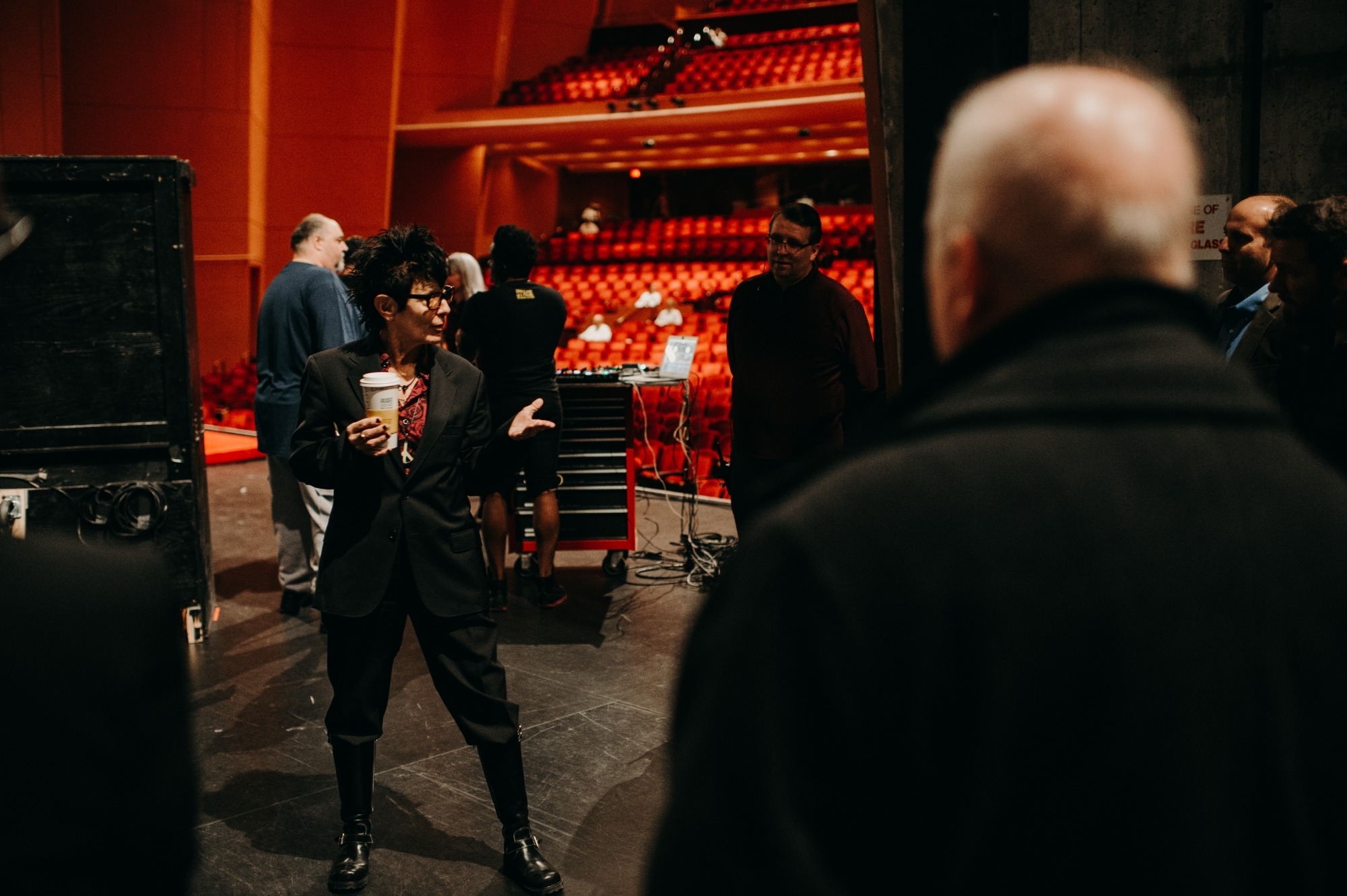 Elizabeth Streb wearing a black suit holding a cup of coffee gesturing to a group of people.