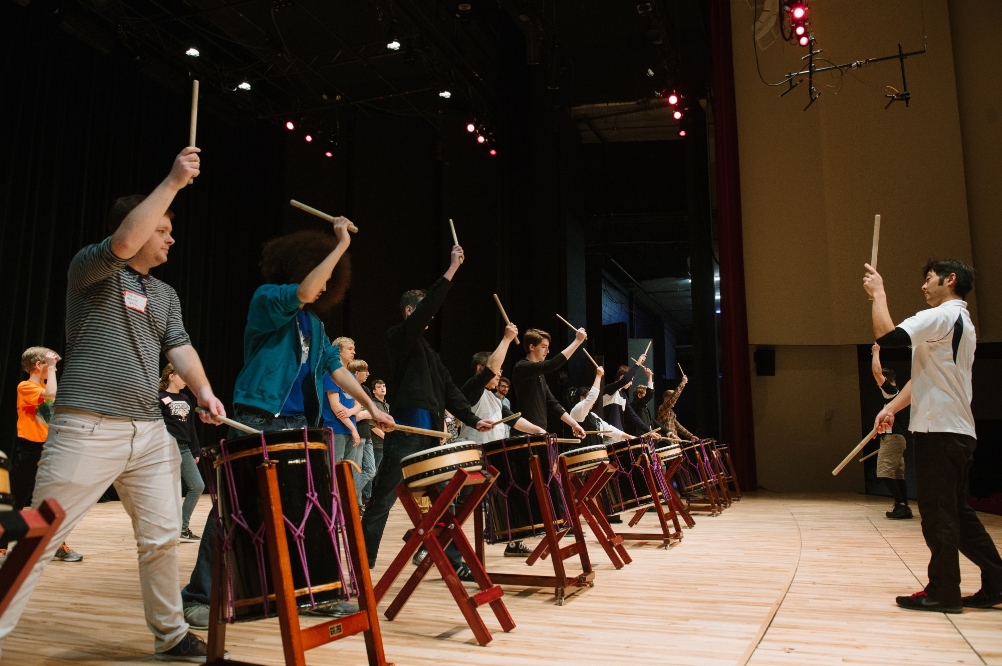 Image of a Taiko drummer leading community members in front of drums with right hands holding a drumstick in the air during a dr