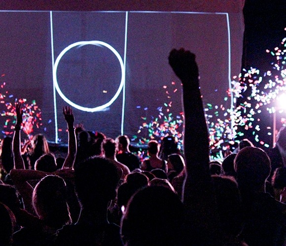 Image from the back of the hall toward the stage showing UNL students in the audience with hands raised and confetti flying in the air during a free Ok Go concert at the Lied Center