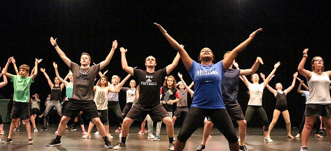 Image of students led by a Matilda performer holding their arms spread apart above their heads during a dance master class