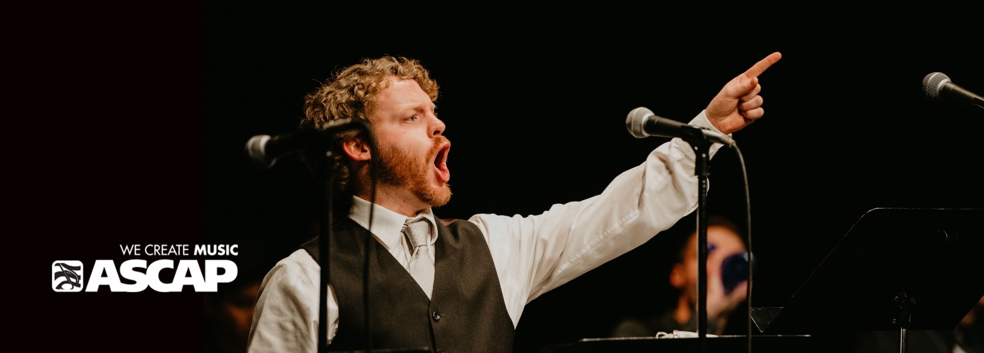 Young man singing behind music stands and microphones against a black background