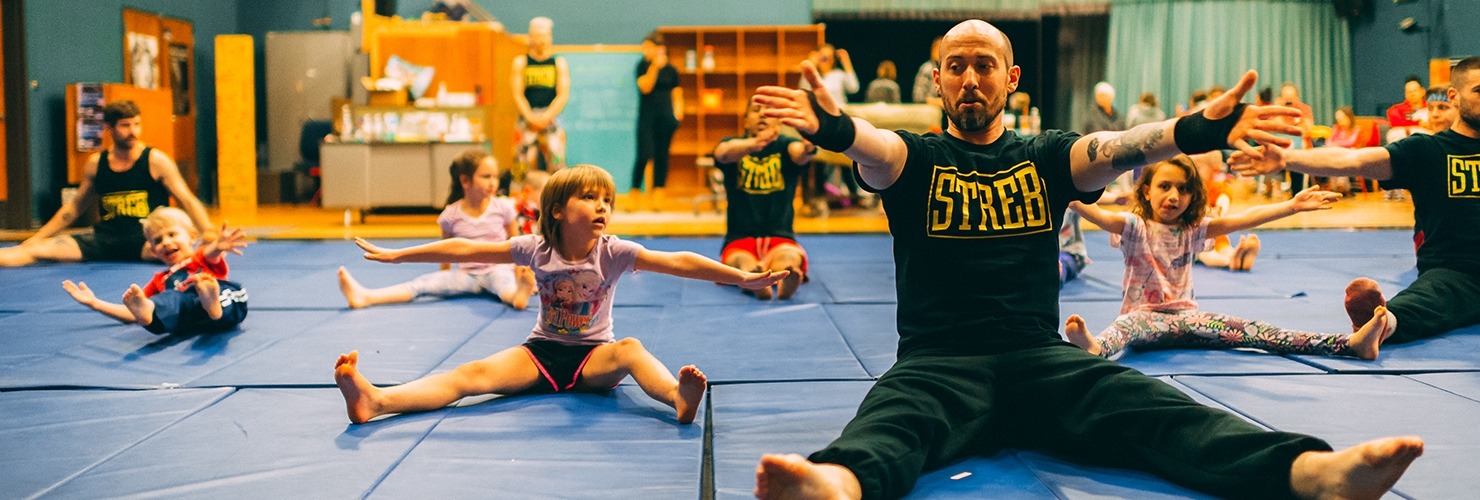 Image of a STREB performer and children sitting on a blue dance mat with arms and legs stretched out forward during a dance master class