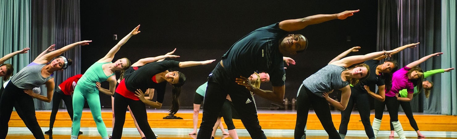 Image of a dancer and students during an Alvin Ailey master class leaning to their left with right arms extended over their heads